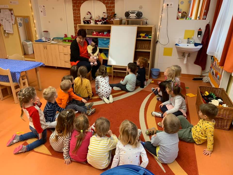 Persona Doll foglalkozás. A pedagógus fogja az ölében a Persona Dollt, előtte gyermekek ülnek a szőnyegen. Figyelik a pedagógust.