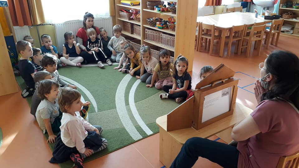 Gyermekek szőnyegen ülve hallgatják az óvodapedagógus meséjét, amit papírszínház módszerrel valósít meg.